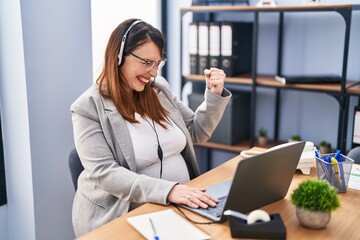 Pregnant woman working at the office wearing operator headset screaming proud, celebrating victory and success very excited with raised arm