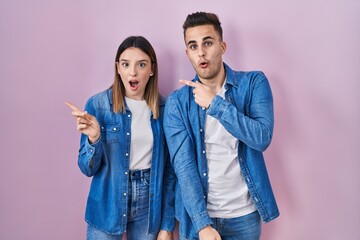 Young hispanic couple standing over pink background surprised pointing with finger to the side, open mouth amazed expression.