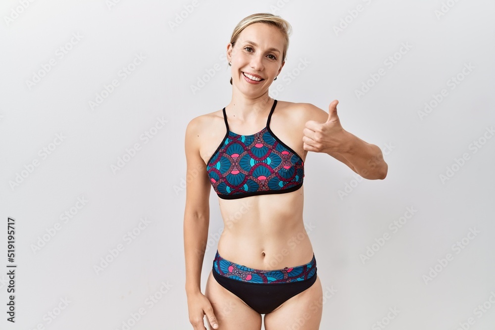 Poster Young blonde woman wearing sporty bikini over isolated background doing happy thumbs up gesture with hand. approving expression looking at the camera showing success.