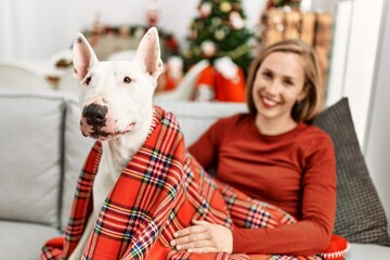 Young caucasian woman smiling confident sitting with dog by christmas tree at home