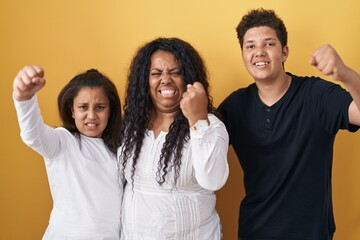 Family of mother, daughter and son standing over yellow background angry and mad raising fist frustrated and furious while shouting with anger. rage and aggressive concept.