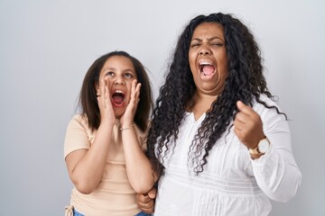 Mother and young daughter standing over white background shouting angry out loud with hands over mouth