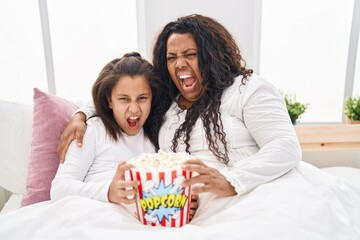 Mother and young daughter eating popcorn in the bed angry and mad screaming frustrated and furious,...