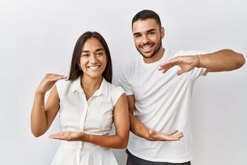 Obraz premium Young interracial couple standing together in love over isolated background gesturing with hands showing big and large size sign, measure symbol. smiling looking at the camera. measuring concept.