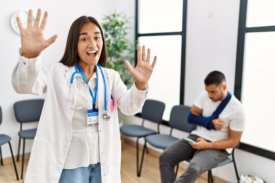 Young asian doctor woman at waiting room with a man with a broken arm showing and pointing up with fingers number ten while smiling confident and happy.