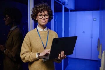 Portrait of female technician in eyeglasses using her laptop while working in data center with...