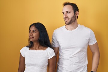 Interracial couple standing over yellow background looking away to side with smile on face, natural expression. laughing confident.