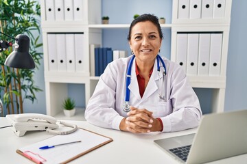 Middle age hispanic woman wearing doctor uniform looking to the camera at clinic