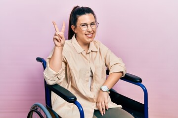 Young hispanic woman sitting on wheelchair smiling looking to the camera showing fingers doing...