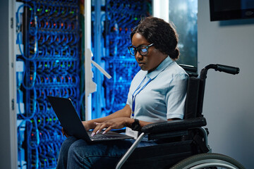 African female IT engineer with disability sitting in wheelchair and typing data on laptop during...