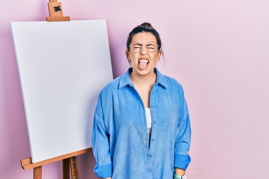 Young hispanic woman standing by painter easel stand sticking tongue out happy with funny expression. emotion concept.