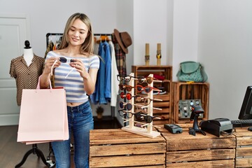 Young chinese woman smiling confident holding shopping bags and sunglasses at clothing store