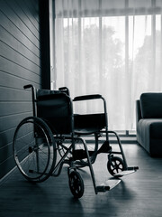 Fototapeta na wymiar Empty black wheelchair near the blue wall and grey with white curtain at glass window in the living room vertical style, waiting for patient services. Lonely wheelchair with nobody in hospital room.