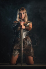 Woman warrior in the armor and with the sword in hands stands on her knees.