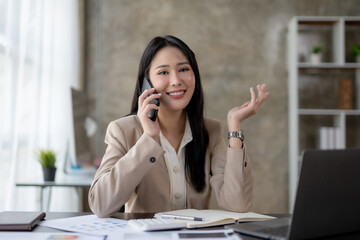 Charming and smiling Asian businesswoman chatting on mobile phone with customer in office.