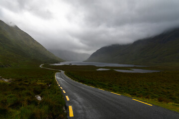 Naklejka premium narrow black-top highway leads through an overcast mountain valley with fog and mist and lakes o nthe valley bottom