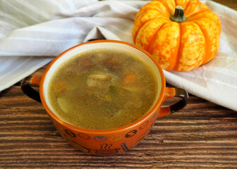 mushroom soup with Polish mushrooms, carrots, potatoes and onions in a brown plate with handles stands on a brown wooden table next to a pumpkin and a gray towel. side view. a bowl of soup