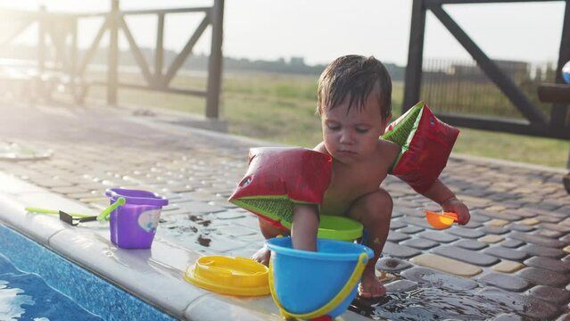 Boy with armbands playing with toys near the pool with clear water on the background of a summer sunset