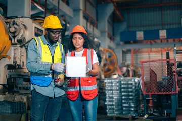 Two professional engineer/worker/technician use clipboard discuss work, walk in steel metal manufacture factory plant industry. Black African American man and woman wear hard hat check quality machine