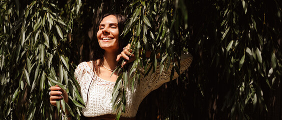 Smiling woman in knitted dress standing near willow trees, banner shot  © Dmytro Hai