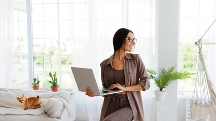 Smiling freelancer in eyeglasses holding laptop near Chihuahua on couch in living room 