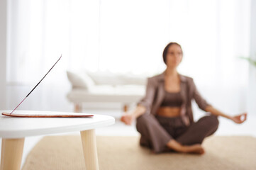 Aromatic stick on table near blurred woman meditating in living room 