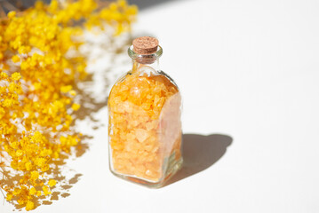 Transparent bottle filled with yellow bath sea salt with wild flowers on white background. Beauty treatment for spa and wellness. Skincare natural cosmetic concept for body care.