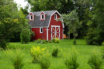 Obraz premium Selective focus view of small red barn with grey shingled roof in field with mixed trees seen in summer in a rural area of Quebec City, Quebec, Canada
