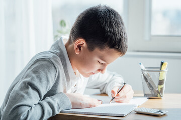 Distance learning online education. Caucasian smile kid boy studying at home, writing in notepad and doing school homework. Thinking child siting at table with notebook. Back to school concept