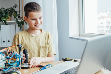 Boy using screwdriver while fixing bolts on a robot. Smart kid looking some tutorial at the laptop computer. Robotics and software engineering for elementary students concept
