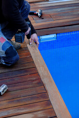 Man wearing working clothes is making a hole on the swimming pool wooden brown deck with the drilling machine. Blue clear water in front of him
