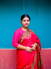 South asian young hindu housewife standing before a tin shed green walled village house, Bangladeshi woman in traditional red dress