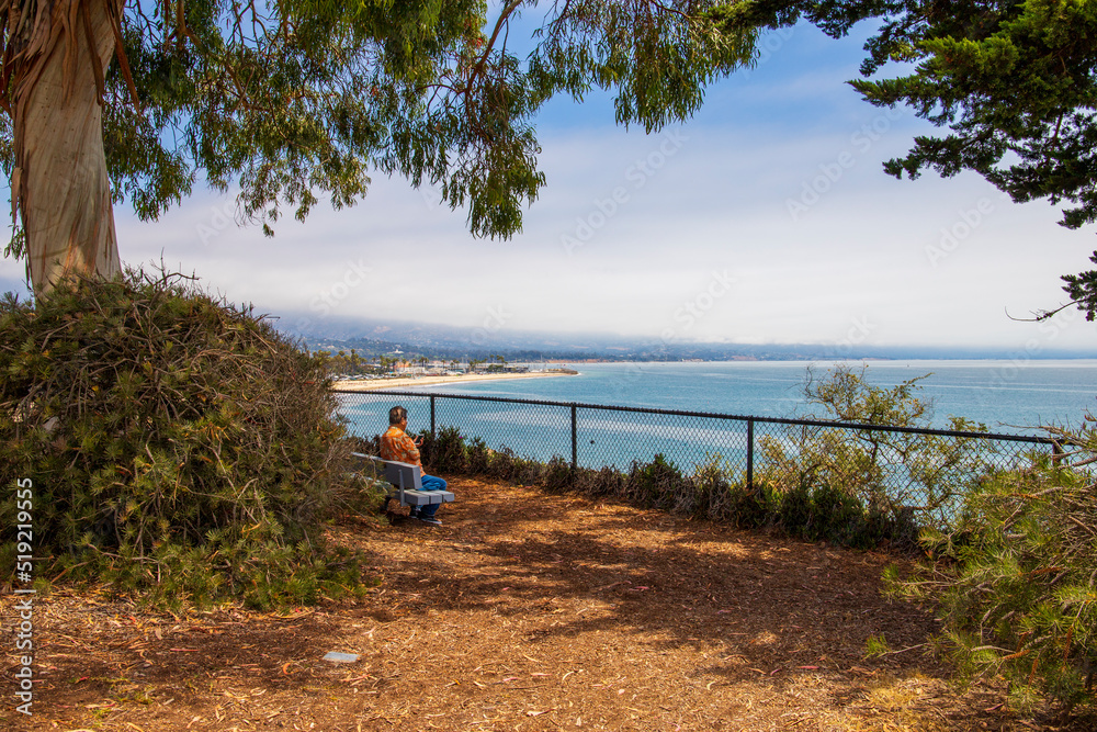 Wall mural an Asian man wearing an orange shirt with one arm sitting on a bench surrounded by blue ocean water and lush green trees, grass and plants with blue sky and clouds at Shoreline Park in Santa Barbara