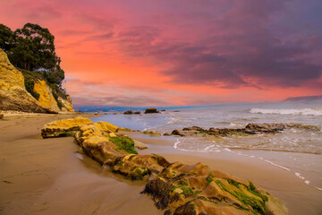 a gorgeous summer landscape at the beach with large rocks in silky brown sand surrounded by blue ocean water and waves rolling into the beach with powerful clouds at sunset at Leadbetter Beach