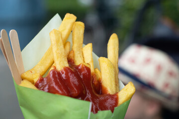 Close up a portion of fries with ketchup in a paper cone with some wooden forks. Fried street food outside, a blurred child's hat in the background.