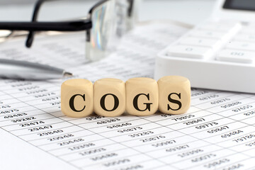 wooden cubes with the word COGS on a financial background with chart, calculator, pen and glasses, business concept.