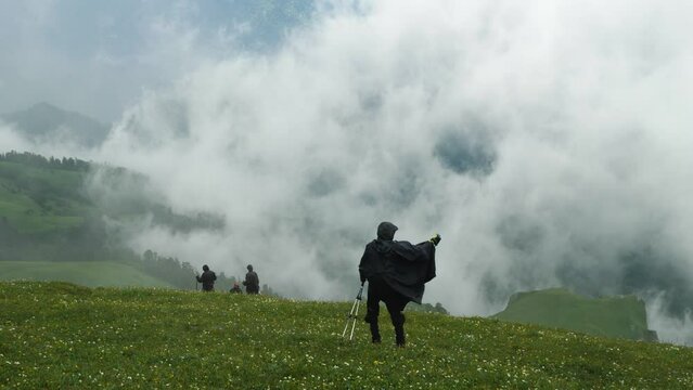A hiker blogger in a black raincoat, trekking poles in hand, descends a grassy slope and shoots himself with an action camera. In the background, the mountain is obscured by clouds and other hikers