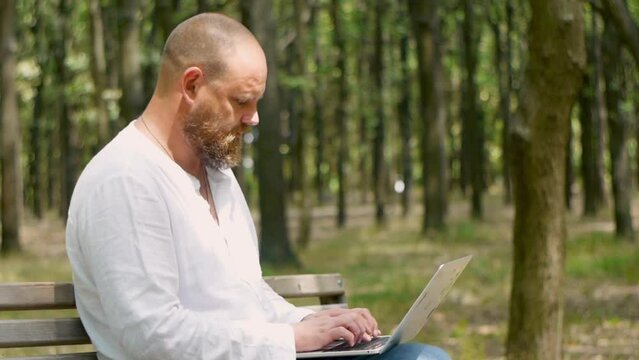 A man with a beard in the park on a bench with a macbook