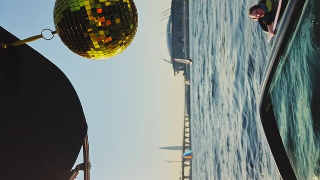 yellow discoball hanging swinging and twirling on a boat behind sunset, around water and bridges, other sailing boats