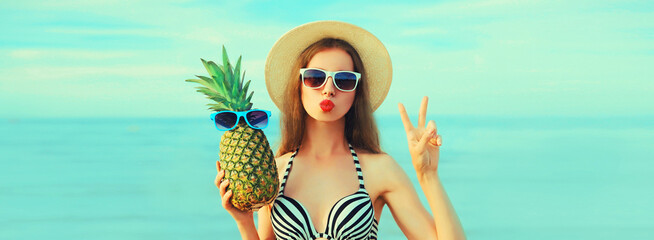 Portrait of happy young woman blowing her lips sends kiss with pineapple in sunglasses, straw hat on the beach on sea background at summer day