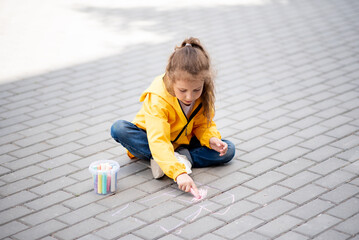 A girl of six years old draws a house on the asphalt with crayons. It's spring and warm outside. The concept of coziness. Everyone needs a home. Childhood. Street.