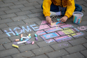 A girl of six years old draws a house on the asphalt with crayons. It's spring and warm outside. The concept of coziness. Everyone needs a home. Childhood. Street.