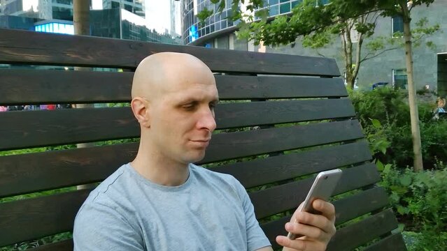 Young man sitting on bench in city park and using mobile phone. Bald guy with smartphone. Man looking carefully at phone screen. Chatting, viewing photos and videos, playing games, Internet surfing