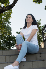 A young brunette woman in a white t-shirt and blue jeans sits on the stairs in the park.