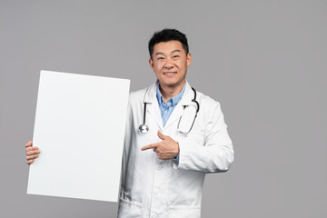 Smiling handsome adult asian male doctor in white coat with stethoscope points finger at banner