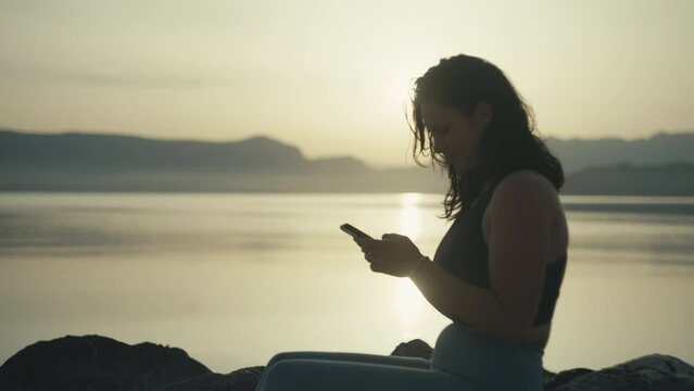 girl sends a message on the phone sitting at sunset by the sea. Woman orders food online on gadget and works in beautiful scenery. High quality 4k footage