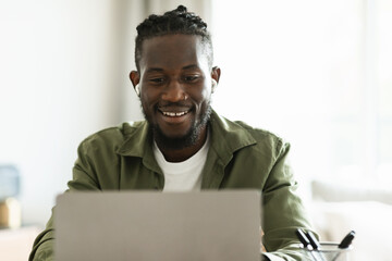 Happy black guy in earphones working on laptop computer at home, enjoying freelance remote job or watching webinar