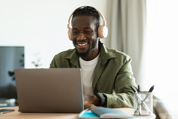 Happy african american man in headset working on laptop computer, sitting at home office and consulting clients online