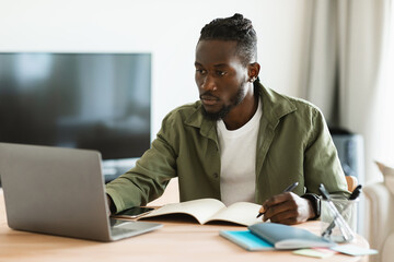 Fototapeta na wymiar Focused african american man using laptop, working remotely and taking notes, having remote meeting from home