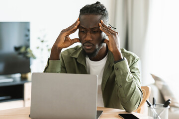 Upset black freelancer man sitting at workdesk at home office, working on laptop and suffering from headache